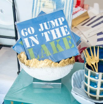 A styled collection of Lake Martin themed home decor and tableware, including blue napkins reading 'GO JUMP IN THE LAKE', a turquoise book, a blue and white checkered cup with gold forks, and a blue speckled rectangular plate.