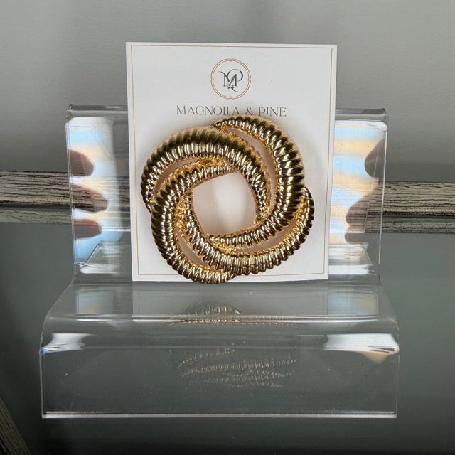 A gold-toned twisted sbrooch displayed on a white Magnolia & Dine brand card, set inside a clear acrylic stand.