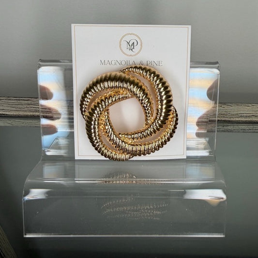 A gold-toned twisted sbrooch displayed on a white Magnolia & Dine brand card, set inside a clear acrylic stand.
