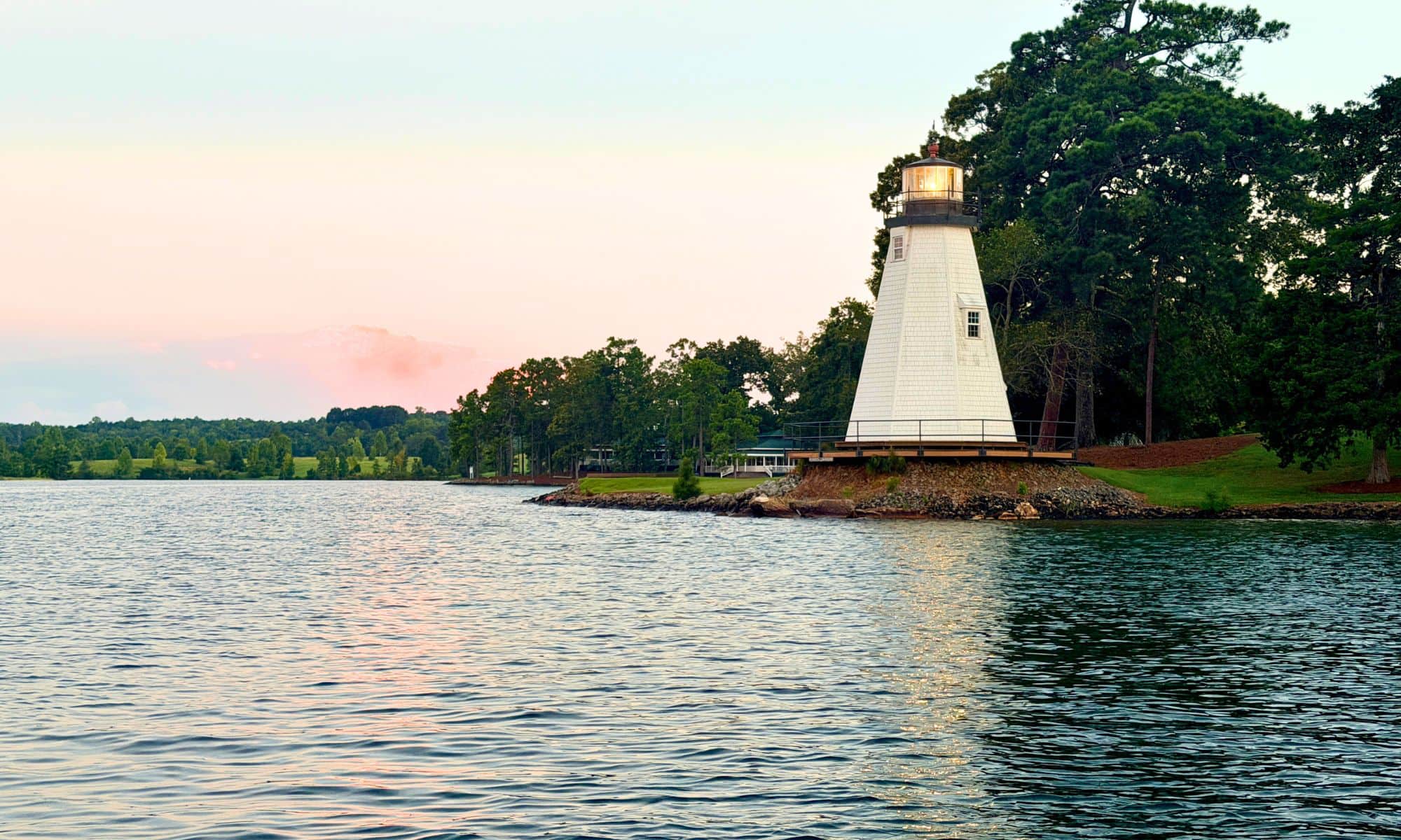 The white lighthouse at Children's Harbor on Lake Martin.  Stands on a grassy shoreline beside calm water, with trees and a soft pink-and-blue sky in the background.