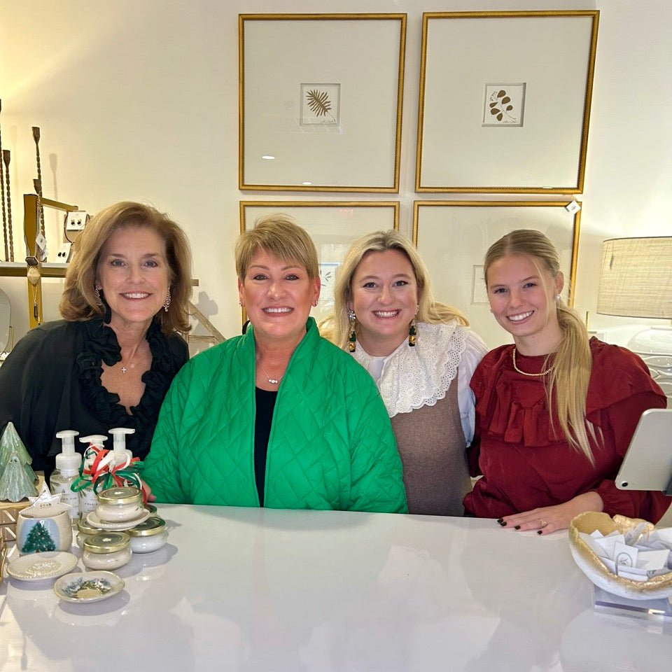 Four women smiling and posing together behind a white counter in a bright, decorated home interior. The center woman wears a bright green shawl over a black top, and the woman on the far right wears a red ruffled top.