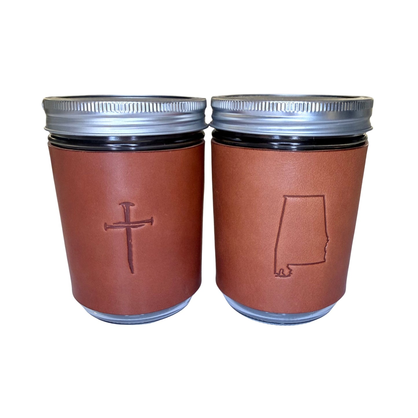 Two brown leather coffee cup sleeves on glass mason jars with silver metal lids, shown against a plain white background. One sleeve features an embossed Christian cross, and the other features an embossed outline of Alabama.