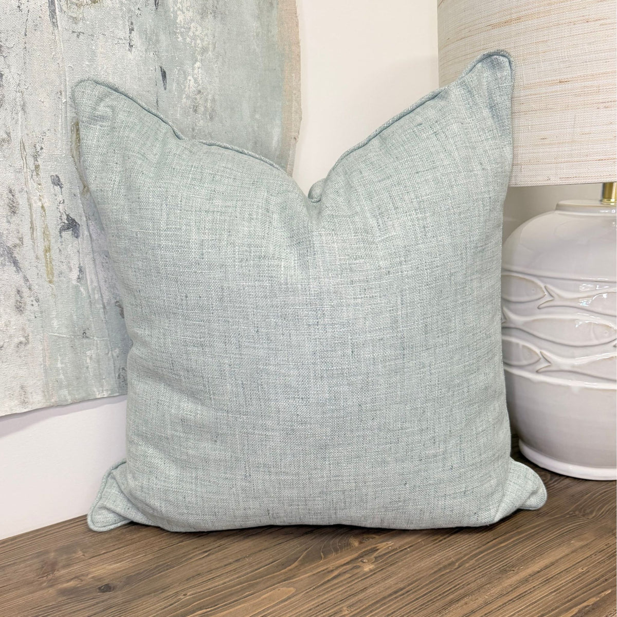 A square throw pillow in a textured light seafoam blue fabric, styled on a wooden floor next to a white ceramic lamp base and a rustic abstract wall art piece.