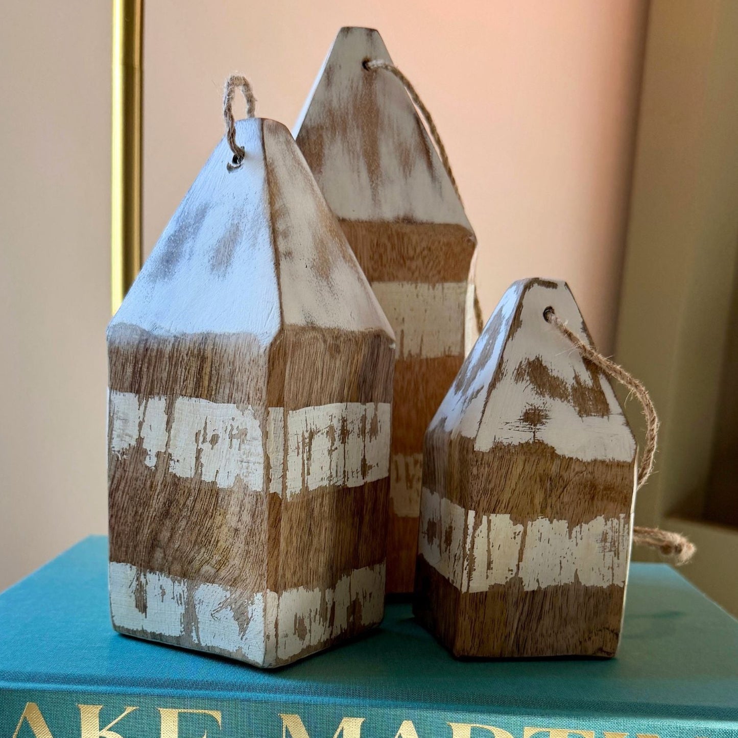 Three rustic wooden house-shaped ornaments with distressed white and natural wood stripes sit on top of a teal book labeled "LAKE MARTIN" on a white pedestal.
