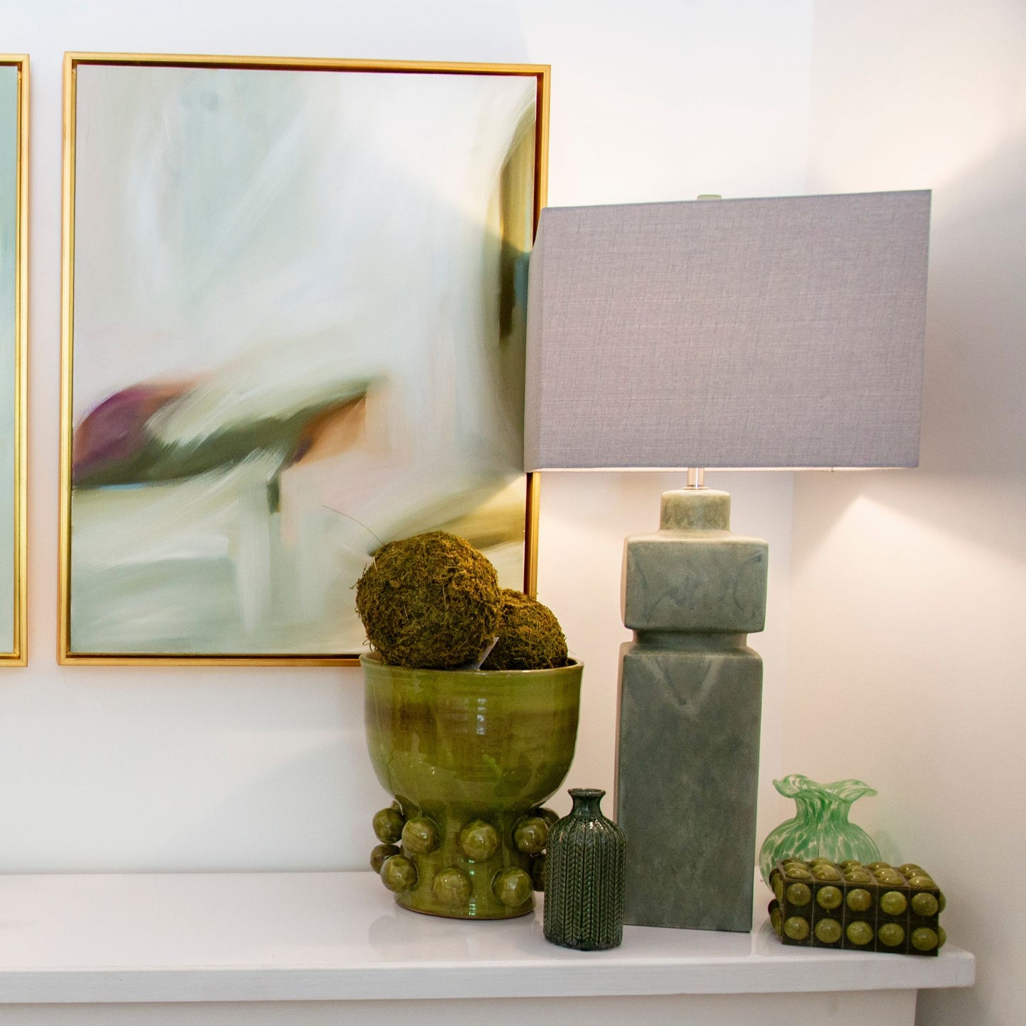 A modern table lamp with a stacked square base in a textured sage green stone-like finish and a gray linen drum shade, styled on a white console table next to a green ceramic bowl and framed abstract wall art.