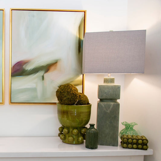 A modern table lamp with a stacked square base in a textured sage green stone-like finish and a gray linen drum shade, styled on a white console table next to a green ceramic bowl and framed abstract wall art.